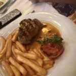 Grilled steak with sauce, crinkle-cut fries, and a grilled tomato garnish on a white plate at a restaurant table.