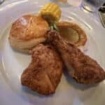 Plate with fried chicken pieces, biscuit, corn on the cob, and mashed potatoes with gravy on a cruise ship dining table.