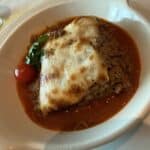 Baked Moussaka with melted cheese, tomato sauce, basil, and a cherry tomato served in a white dish on a tablecloth.