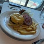 Cheeseburger with lettuce, tomato, onion, and French fries served on a white plate by a window with ocean views on a cruise ship deck