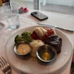 Surf and turf meal with lobster tail, steak, broccoli, mashed potatoes, and sauces on a white plate by a window with ocean views.