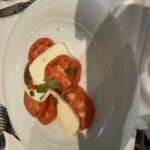 Caprese salad with sliced tomatoes, fresh mozzarella, basil, and pesto drizzle on a white plate at a restaurant table setting