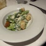 Caesar salad with romaine lettuce, croutons, grated cheese, and creamy dressing served on a white plate at a restaurant table.