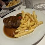 Grilled steak with peppercorn sauce, French fries, and a grilled tomato served on a white plate at a restaurant table.