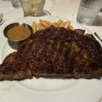 Grilled steak with French fries and a side of brown gravy sauce served on a white plate at a restaurant table.