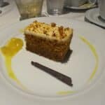 Slice of carrot cake with cream cheese frosting, chocolate garnish, and mango sauce on a white plate at a cruise ship dining table.