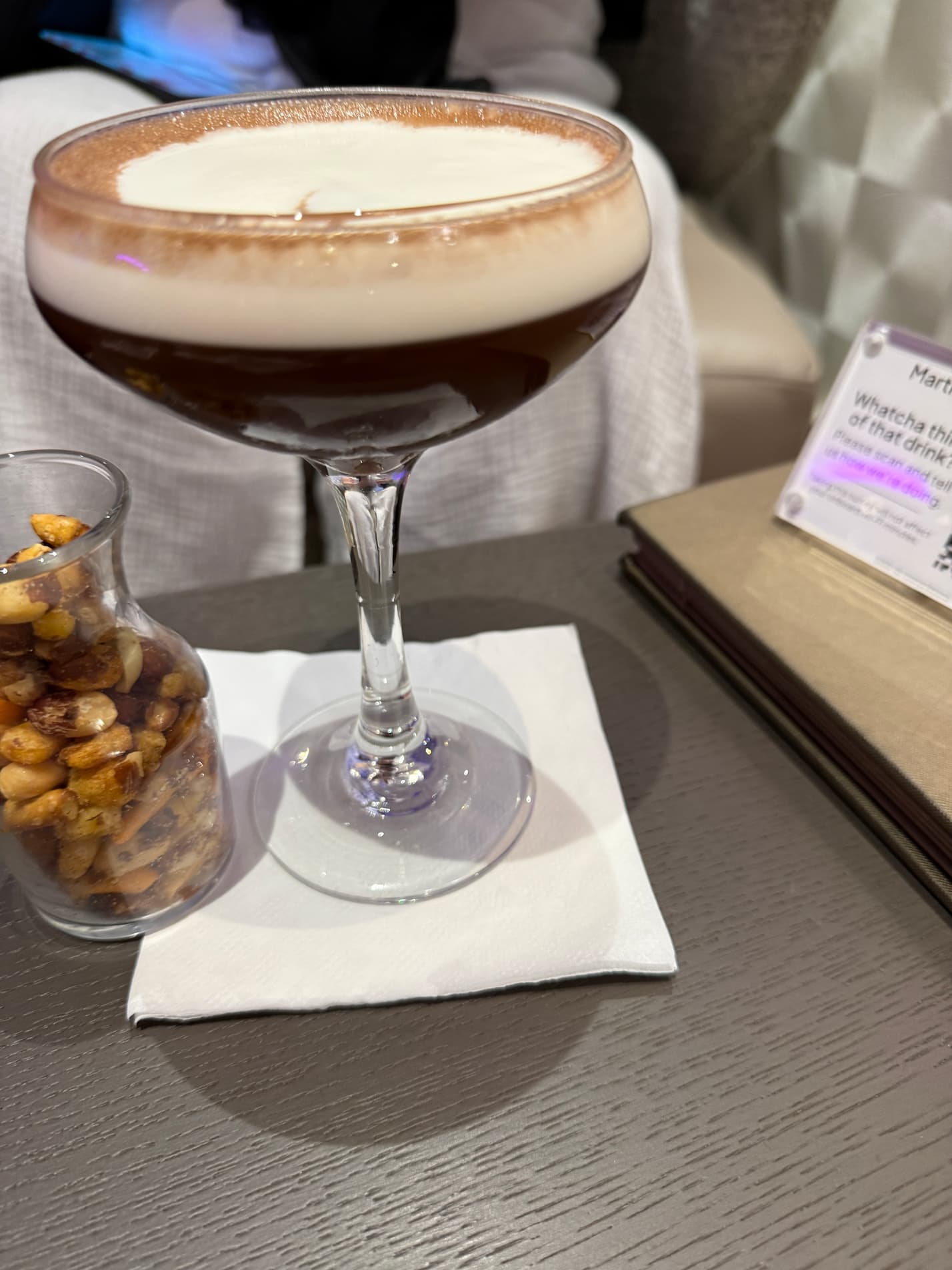 What Drinks Are Included on a Cruise? Free vs. Paid Drinks 3 Espresso martini cocktail on a napkin next to a small jar of mixed nuts on a table in a lounge setting.
