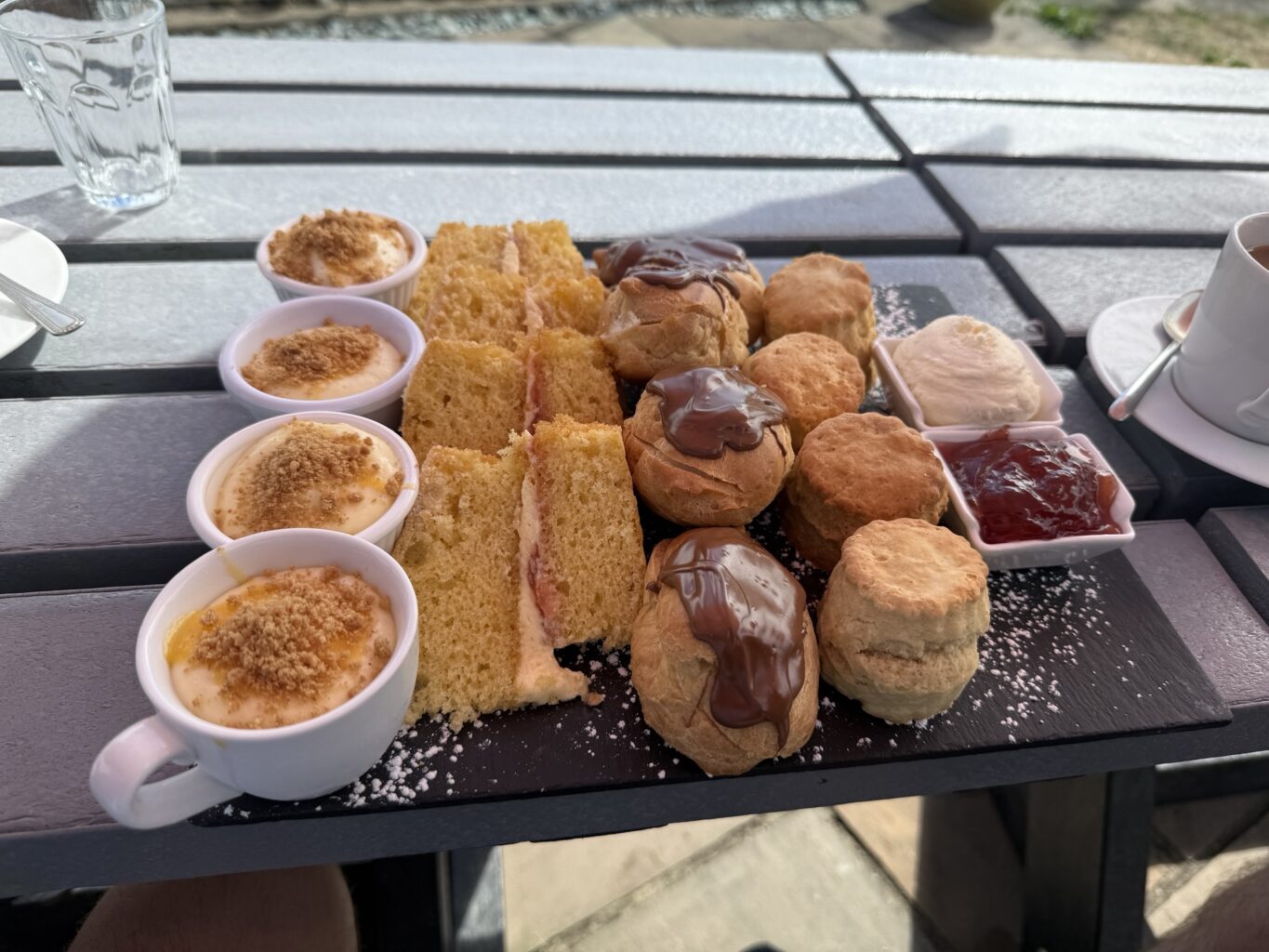 afternoon-tea-dessert-platter-with-scones-cakes-and-cream.jpeg Assorted afternoon tea treats including sponge cake, chocolate eclairs, scones with clotted cream and jam, and custard cups on an outdoor table.