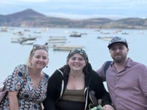 Nicole-Lilly-TJ-Ty-Coch-Inn.jpeg Three people smiling near a calm harbor with boats and hills in the background during a family vacation by the water
