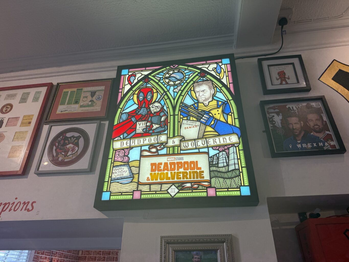 deadpool-and-wolverine-stained-glass-wall-art.jpeg Stained glass-style wall art featuring Deadpool and Wolverine with framed comic and movie memorabilia around it.