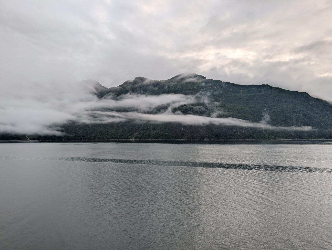 foggy-mountain-over-calm-water.jpg Calm ocean waters with low clouds drifting around a forested mountain under a cloudy sky, ideal for a cruise vacation.